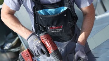 Ein Handwerker in Arbeitskleidung benutzt ein Elektrowerkzeug, um an einem Dach zu arbeiten, während die Sonne scheint.