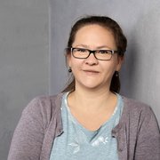 Eine Frau mit Brille lächelt freundlich, während sie an einer grauen Wand lehnt, in einem lässigen Outfit.