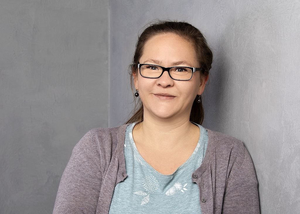 Eine Frau mit Brille lächelt freundlich, während sie an einer grauen Wand lehnt, in einem lässigen Outfit.
