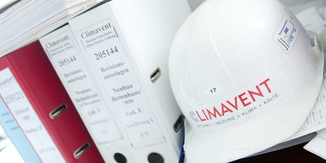 Ein weißer Bauhelm mit dem Logo von Climavent liegt auf einem Stapel von Dokumenten und Aktenordnern.