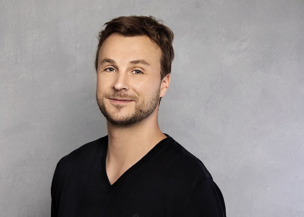 Der Mann hat kurzes, braunes Haar und einen gepflegten Bart, während er freundlich in die Kamera schaut.