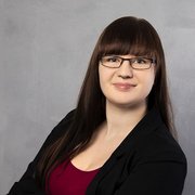 Die Person trägt ein schwarzes Blazer-Jackett und ein rotes Oberteil, während sie selbstbewusst in die Kamera schaut.