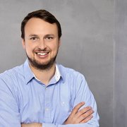 Der Mann mit einem freundlichen Lächeln hat braune Haare und trägt ein gestreiftes Hemd, während er entspannt posiert.