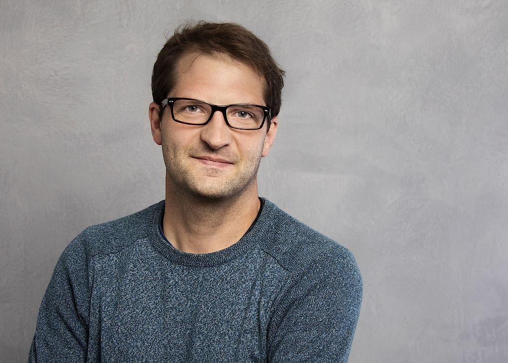 Der Mann mit Brille hat einen freundlichen Ausdruck und steht mit verschränkten Armen vor einem neutralen Hintergrund.