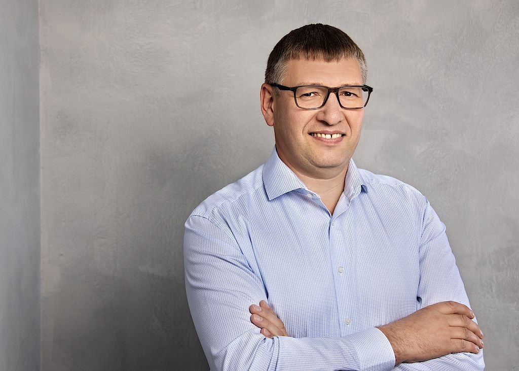 Der Mann hat die Arme verschränkt und zeigt ein freundliches Lächeln, umgeben von einer neutralen, grauen Kulisse.