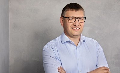 Der Mann hat die Arme verschränkt und zeigt ein freundliches Lächeln, umgeben von einer neutralen, grauen Kulisse.