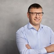 Der Mann hat die Arme verschränkt und zeigt ein freundliches Lächeln, umgeben von einer neutralen, grauen Kulisse.